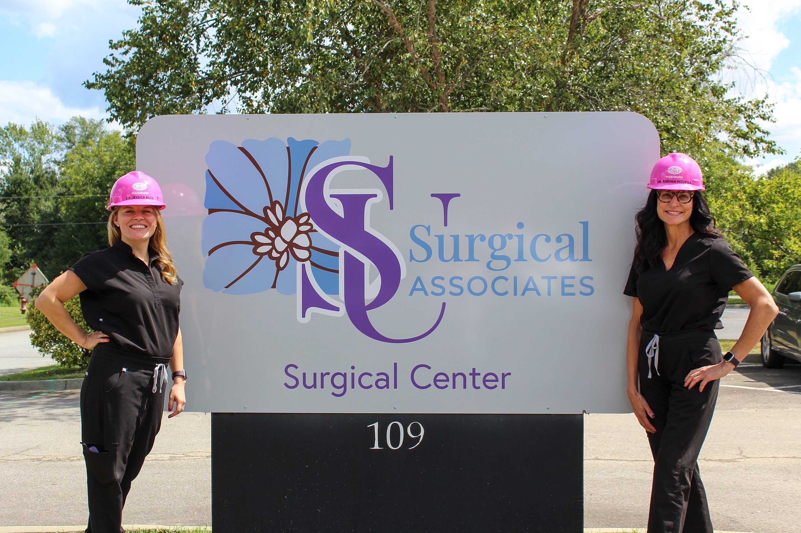 Two women in surgical attire beside a sign.